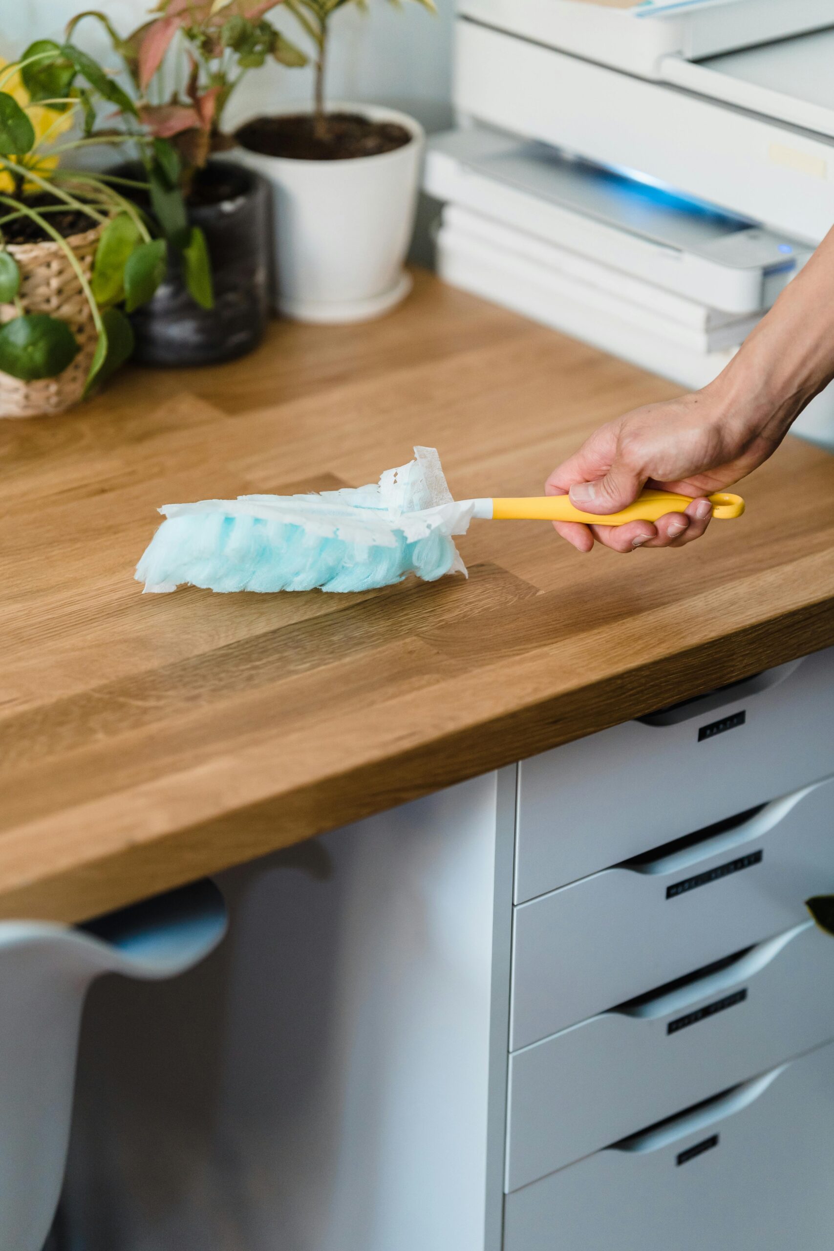 Commercial A hand using a feather duster to clean a wooden desk, surrounded by plants and office items.